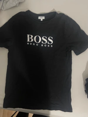Svart Hugo Boss t-shirt med logga - Snygg svart t-shirt från Hugo Boss med stor vit logga på bröstet. Klassisk rund halsringning och korta ärmar. Tillverkad i mjuk bomull som känns skön mot huden. Perfekt för dig som gillar stilrena och tidlösa plagg.