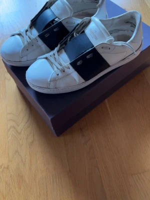 Valentino vita sneakers med svart band - Snygga vita sneakers från Valentino med ett brett svart band över vristen och diskreta detaljer. Skorna har klassisk låg profil och är tillverkade i läder med gummisula. Perfekta för dig som gillar exklusiv och stilren design.