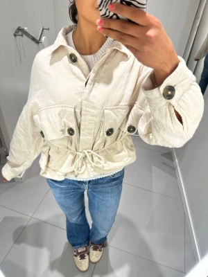 Beige manchester overshirt med knyt - Säljer en beige overshirt i manchester med stora knappar, knyt i midjan och två stora bröstfickor. Jackan har en avslappnad passform och långa ärmar med knappdetaljer vid mudden. Perfekt för lager på lager och en chill look.