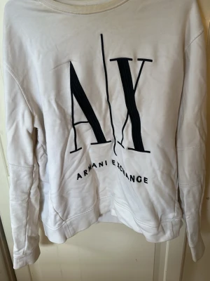Vit sweatshirt från Armani Exchange - Snygg vit sweatshirt från Armani Exchange med stor svart AX-logga framtill. Tröjan har rund halsringning och ribbade muddar vid ärmslut och nederkant. Perfekt för en clean och trendig look.