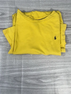 Gul t-shirt från Polo Ralph Lauren - Gul t-shirt från Polo Ralph Lauren med klassisk passform och liten svart logga broderad på bröstet. Tillverkad i mjuk bomull och har rund halsringning samt korta ärmar. Perfekt för dig som gillar stilrena och färgglada plagg.