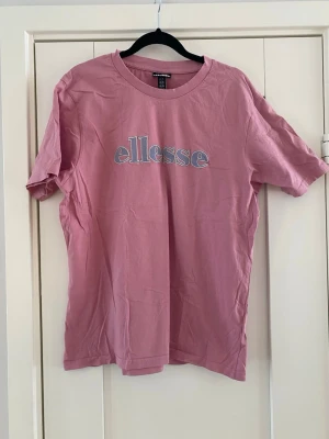 Rosa ellesse t-shirt med broderad logga - Snygg rosa t-shirt från ellesse med broderad logga i vitt och ljusblått på bröstet. Klassisk rund halsringning och korta ärmar. Tillverkad i mjuk bomull som känns skön mot huden. Perfekt för en avslappnad och trendig look. Sparsamt använd och i bra begagnat skick   Mått Över bröstet: ca 54 cm Längd från en axel och ner: ca 67 cm  100% bomull  Kommer från ett djur och rökfritt hem