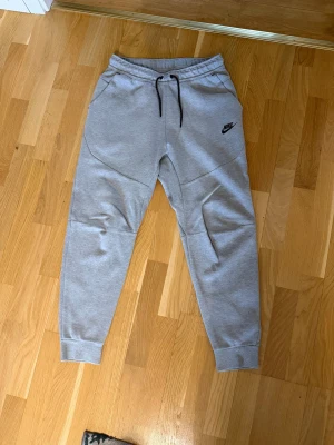 Gråa Nike Tech Fleece mjukisbyxor - Säljer ett par gråa Nike Tech Fleece mjukisbyxor med svarta dragsnören och logga på vänster ben. Byxorna har ribbade muddar, fickor fram och en snygg zipficka bak. Perfekta för chill eller streetwear-stil.