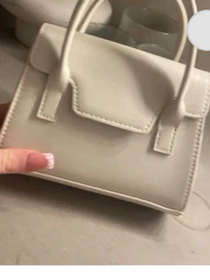 Vit minihandväska med axelrem - Snygg vit minihandväska i slätt konstläder med två handtag och avtagbar, justerbar axelrem. Väskan har en stilren, fyrkantig form och lock med sömdetaljer. Perfekt för dig som gillar minimalistisk och trendig design.