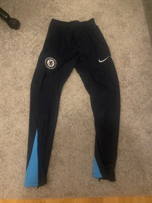 Chelsea FC träningsbyxor Nike - Mörkblå träningsbyxor från Nike med Chelsea FC-logga på låret och vit Nike Swoosh. Byxorna har smal passform, elastisk midja och blå detaljer vid bensluten. Perfekta för fotbollsträning eller chill. Tillverkade i ett lätt och funktionellt material.