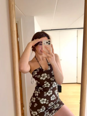 Brun blommig halterneckklänning - Säljer en brun kort klänning med  blommor och halterneck. Klänningen har en tight passform och är ärmlös, så snygg till sommaren med tan 😍  !