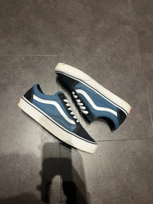 Blå Vans Old Skool sneakers - Klassiska Vans Old Skool sneakers i blått och svart med vit sula och vit sidostripes. Ovandelen är i canvas och mocka, med vita skosnören och låg profil. Perfekta för en avslappnad streetwear-look.