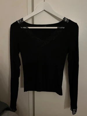 Svart långärmad topp med spets - Snygg svart långärmad topp med v-ringning och spetsdetaljer vid halsringning och ärmslut. Perfekt att styla med jeans eller kjol för en trendig look.