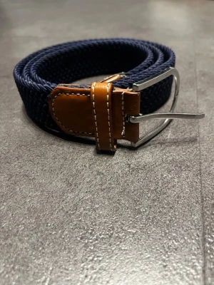 Marinblått flätat bälte med skinn - Snyggt marinblått flätat bälte med detaljer i brunt skinn och silverfärgat spänne. Perfekt accessoar för att lyfta din outfit med en klassisk touch. Bältet har en flexibel design och passar till många olika stilar.