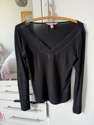 Svart långärmad topp med spets - Snygg svart långärmad topp från hunkemöller med djup v-ringning och spetsdetaljer längs urringningen. Figurnära passform som framhäver siluetten. Det finns ett längst ut på ärmen (se bild) men det är inget som är tydligt när man har den på sig.