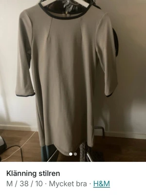 Beige klänning med svarta detaljer H&M - Stilren beige klänning från H&M med svarta kontrastdetaljer vid halsringning och ärmslut. Klänningen har trekvartsärmar och rak passform, perfekt för en enkel och snygg look. Materialet känns mjukt och följsamt.