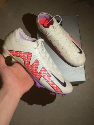 Nike vita Mercurial Vapor 16 Elite  - Säljer ett par Nike Mercurial fotbollsskor i vitt med lila och röda detaljer. Skorna har en modern, strömlinjeformad design med svart Nike-logga och mönstrad sula. Ovandelen är tillverkad i syntetmaterial för bra bollkontroll och grepp på planen.