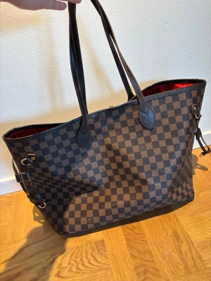 Totebag med Damiet Ebene mönster shoppingväska - Säljer en Totebag i Damier Ebene-mönster. Väskan har brunt rutigt mönster, mörkbruna handtag och detaljer samt röd textilinsida med tryck. Rymlig modell med innerficka och avtagbar liten väska. Perfekt för dig som gillar lyxig och tidlös stil.