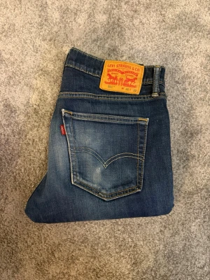 Levi's 511 blå jeans herr - Säljer ett par klassiska blå Levi's 511 jeans med snygg tvätt och slitningar. Jeansen har raka ben, fem fickor och den ikoniska röda Levi's-lappen på bakfickan. Tillverkade i bomull med en skön passform som funkar till det mesta.