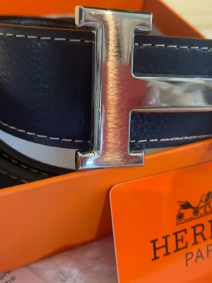 blått skärp med H-spänne - Snyggt blått skärp med ikoniskt H-format metallspänne. Skärpet har vita kontrastsömmar. Perfekt accessoar för att lyfta din outfit.