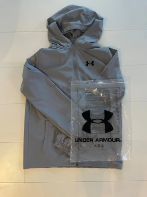 Grå windbreaker från Under Armour - Snygg grå vindjacka från Under Armour med huva och dragkedja framtill. Jackan har två sidofickor och elastiska muddar vid ärmsluten. Tillverkad i lätt och tåligt material, perfekt för blåsiga dagar. Stor logga på bröstet för sportig look. Använd 1 gång, mycket bra skick! Pris kan diskuteras☺️ 
