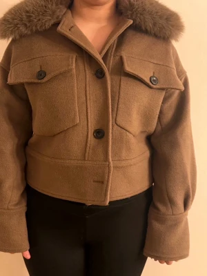 Meotin kappa  - Kort brun kappa med bred krage i fuskpäls, stora bröstfickor med lock och knappar framtill. Rak passform och långa ärmar med breda muddar. Perfekt för kyliga dagar och ger en trendig look.