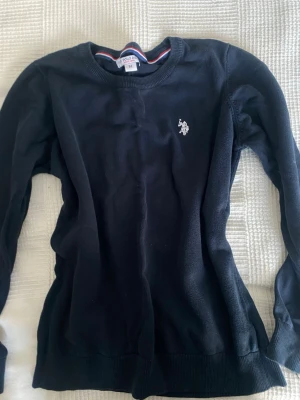 Mörkblå stickad tröja US Polo Assn - Snygg mörkblå stickad tröja från US Polo Assn med rund halsringning och vit broderad logga på bröstet. Tröjan har ribbade muddar vid ärmslut och nederkant samt diskreta ränder i kragen. Perfekt till en clean och klassisk stil.