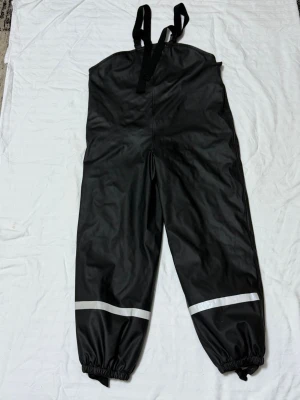 Svarta täckbyxor från Mywear Active - Svarta täckbyxor med hängslen från Mywear Active. Byxorna har resår vid bensluten och reflexband runt benen för extra synlighet. Insidan är fodrad med mjuk grön fleece som håller dig varm under kalla dagar. Perfekta för vinterns alla äventyr.