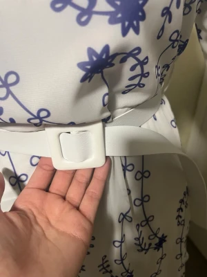 Vit klänning med blått blommönster och bälte - Säljer en vit klänning med blått blommönster och ett brett vitt bälte med fyrkantigt spänne i midjan. Klänningen har en flowy passform och är tillverkad i ett lätt tyg som känns som polyester. Perfekt för dig som gillar unika detaljer och mönster! Finns två av den  , köpt utomlands, priset kan diskuteras .  Aldrig använt !!!  Passar I s och xs