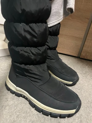 Svarta moon boots med grov sula - Säljer ett par svarta moon boots med puffig design och grov, vit och svart sula. Skorna har dragkedja på sidan och är tillverkade i ett vattenavvisande syntetmaterial. Perfekta för kalla dagar när du vill hålla fötterna varma och torra.