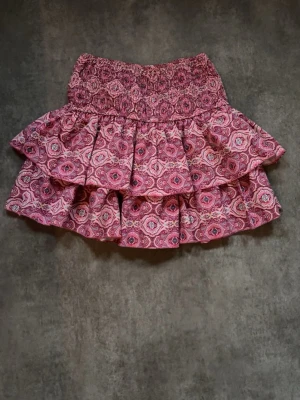 Rosa volangkjol från MEET x TIGER - Supersöt kort volangkjol från MEET x TIGER i rosa med mönster och bred smock i midjan. Kjolen har dubbla lager volanger och ett bohemiskt print som gör den perfekt för vår och sommar. Lätt och luftig känsla, passar till både sneakers och sandaler. Det är inbyggda shorts i kjolen!