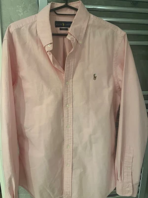 Rosa skjorta från Ralph Lauren - Klassisk rosa skjorta från Ralph Lauren med broderad logga på bröstet. Skjortan har knappar framtill, button-down krage och långa ärmar. Tillverkad i mjuk bomull som känns skön mot huden. Perfekt för en clean och stilren look.