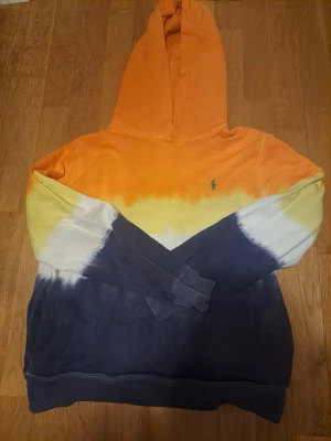 Färgglad hoodie från Polo Ralph Lauren - Säljer en riktigt snygg hoodie från Polo Ralph Lauren med huva. Hoodien har en cool tie-dye design i orange, gul, vit och mörkblå. Liten grön logga broderad på bröstet. Perfekt för dig som vill sticka ut med färg och stil. Mjuk bomullskvalitet.