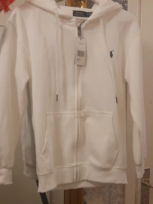 Vit hoodie från Ralph Lauren - Säljer en stilren vit hoodie från Ralph Lauren med dragkedja framtill, två stora fickor och klassisk huva med dragsko. Ikoniska Polo-loggan broderad på bröstet. Etiketter kvar. Oanvänd 
