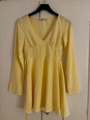 Ljusgul omlottklänning med v-ringning - Söt ljusgul kortklänning med v-ringning och långa ärmar. Klänningen har omlottdetalj framtill och knytband i midjan för en snygg passform. Tillverkad i ett mjukt bomullsliknande material som känns bekvämt mot huden.