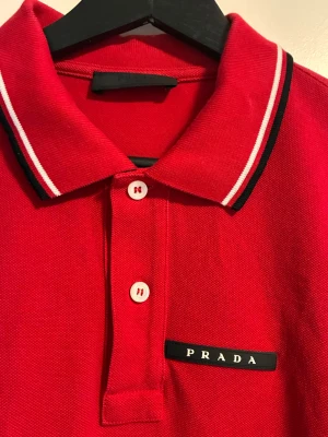 Röd pikétröja från Prada - Snygg röd pikétröja från Prada med klassisk krage och två vita knappar. Kragen och ärmsluten har vita och svarta ränder som ger en sportig touch. Diskret Prada-logga på bröstet. Perfekt till jeans eller shorts.
