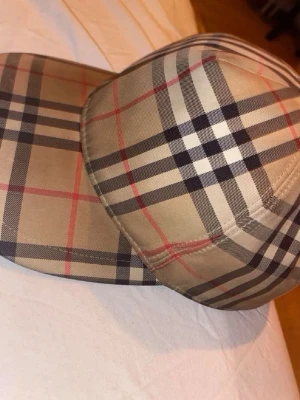 Burberry keps - Snygg keps från Burberry med det klassiska rutmönstret i svart, vitt och rött. Justerbar baktill med svart plastspänne och en patch med texten 'BURBERRY LONDON ENGLAND'. Perfekt accessoar för en trendig streetwear-look. Pris kan diskuteras vid snabb affär
