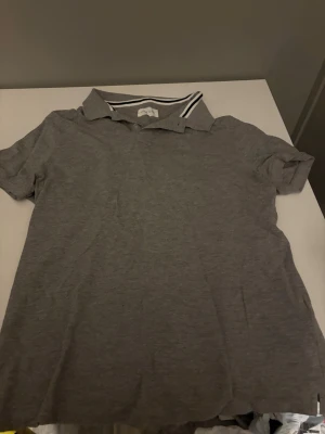 Grå pikétopp med krage - En enkel grå pikétopp med korta ärmar och klassisk krage med vit och svart randig detalj på insidan. Perfekt till jeans eller shorts för en avslappnad look.