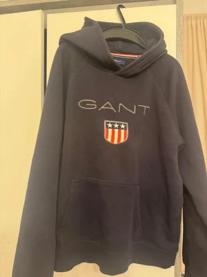 Mörkblå hoodie från GANT med logga - Snygg mörkblå hoodie från GANT med broderad logga och amerikanskinspirerat emblem på bröstet. Klassisk känguruficka framtill och huva. Perfekt för en avslappnad och sportig stil. Mjuk bomullsblandning för extra komfort.