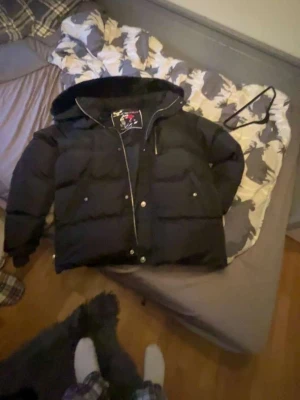 Svart dunjacka från Moose Knuckles - Säljer en svart dunjacka från Moose Knuckles med stor huva och pälskant. Jackan har silverfärgade detaljer, dragkedja och tryckknappar samt den ikoniska loggan på ärmen. Perfekt för kalla vinterdagar och riktigt snygg streetstil.