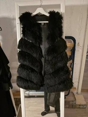 Svart fluffig pälsväst med krage - Snygg svart pälsväst från Rockandblue med fluffig fuskpäls och bred krage. Västen har ett coolt, randigt mönster i pälsen och är ärmlös. Perfekt för att lyfta din outfit med en trendig touch.