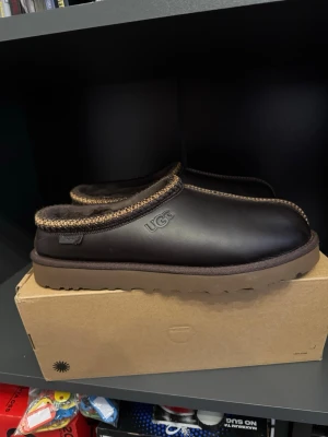 Mörkbruna UGG tofflor i skinn - Säljer ett par mörkbruna tofflor från UGG i skinn med mjuk innersida och grov sula. Tofflorna har dekorativt mönstrad kant i brunt och beige samt UGG-logga på sidan. Perfekta för att hålla fötterna varma hemma.