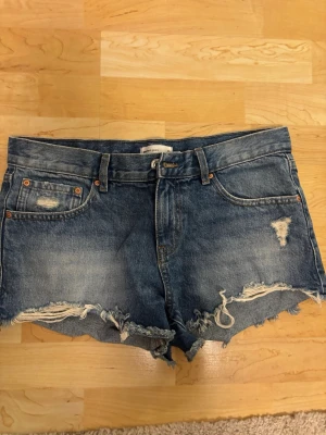 Blå slitna jeansshorts med fransar - Säljer ett par blå jeansshorts med råa, fransiga kanter och slitningar framtill. Klassisk femficksmodell med knapp och dragkedja. Perfekta för en avslappnad och trendig look. Materialet är denim i bomull.