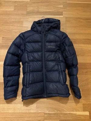 Mörkblå dunjacka från Peak Performance - Snygg mörkblå dunjacka från Peak Performance med huva och dragkedja framtill. Jackan har en puffig design med breda kanaler och diskret logga på bröstet. Perfekt för kalla vinterdagar och riktigt varm tack vare dunfyllningen.