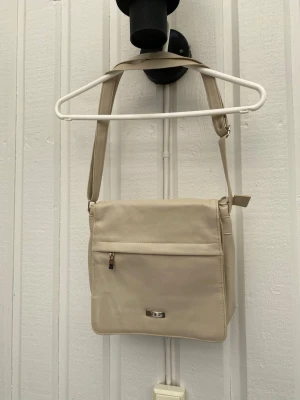 Beige axelväska från D&S - Snygg beige axelväska från D&S i fyrkantig modell. Väskan har justerbar axelrem, olika fack. Gammal väska som legat i ett förråd senaste åren. Några gula fläckar inuti medans vissa fack är i väldigt fint skick. Fler bilder vid förfrågan. Färgen är svår att fånga på bild. Djur och rökfritt hem! 