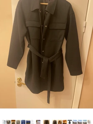 Svart lång cardiganjacka med bälte - Stilren svart cardiganjacka med knappar framtill, två stora bröstfickor och ett matchande midjebälte. Jackan har lång ärm och klassisk krage, perfekt för lager på lager. Materialet ser ut att vara tjockt och mjukt, troligen ullblandning.