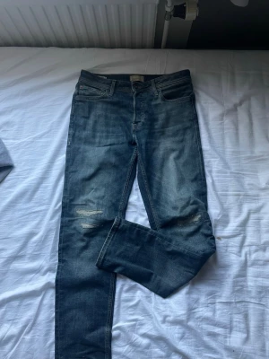 Blå slitna skinny jeans från Jack & Jones - Säljer ett par blå slim fit jeans från Jack & Jones med slitningar över knäna. Jeansen har en snygg tvättad look och är tillverkade i mjuk denim med stretch för extra komfort. Perfekta för en avslappnad och trendig stil. Kom gärna med frågor😊✌🏼