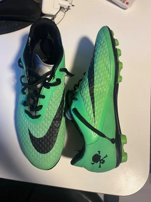 Gröna Nike fotbollsskor med svart sula - Säljer mina Nike hypervenom. Stl 44. Snygga gröna fotbollsskor från Nike med svart sula och svart snörning. Skorna har en texturerad ovansida för bättre bollkontroll och en stor svart Nike-logga på sidan. På hälen finns en dödskalle-detalj. Perfekta för spel på gräsplan.