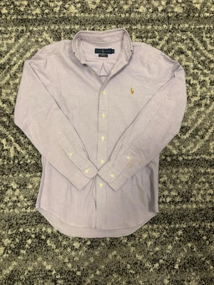 Ljuslila skjorta från Ralph Lauren - Klassisk ljuslila skjorta från Ralph Lauren med button-down krage och broderad logga på bröstet. Skjortan har långa ärmar och vita knappar framtill. Tillverkad i bomull för en bekväm och stilren look. Skjortan är i storlek S! Hör av er vid eventuella frågor eller funderingar!!