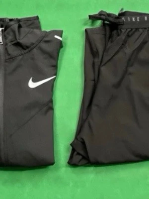 Svart träningsset från Nike - Snyggt svart träningsset från Nike med matchande jacka och byxor. Jackan har huva, dragkedja och vita Nike-loggor på bröstet och axlarna. Byxorna har resår i midjan och diskret Nike-märke. Perfekt för träning eller chill.