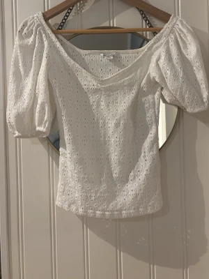 Vit broderad blus med puffärm - Söt vit blus med broderade detaljer och puffärmar. Toppen har v-ringning och är tillverkad i bomull med spetsliknande struktur. Perfekt för dig som gillar romantisk och feminin stil. Passar till både jeans och kjol.