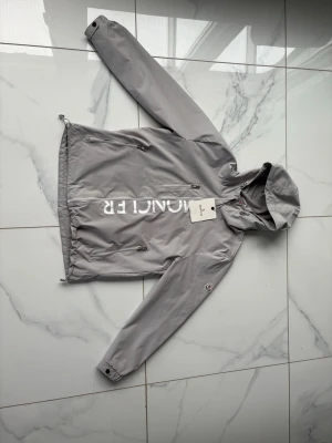 Grå vindjacka från Moncler - Snygg grå vindjacka från Moncler med huva och dragkedja framtill. Jackan har vita Moncler-tryck på bröstet och ett broderat logomärke på ärmen. Praktiska fickor med dragkedja och justerbar nederkant. Perfekt för blåsiga dagar och streetwear-stil.