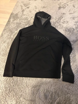 Svart hoodie från Hugo Boss - Snygg svart hoodie från Hugo Boss med stor logga på bröstet. Klassisk huva och magficka framtill. Tillverkad i mjukt material som känns skönt mot huden. Perfekt för en avslappnad och stilren look.