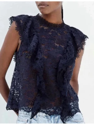 Mörkblå spetsblus med volanger - Trendig mörkblå topp i genombruten spets med volangdetaljer framtill och vid ärmslut. Ärmlös modell med rund halsringning och lätt utsvängd passform. Perfekt att styla med jeans eller kjol för en cool look.