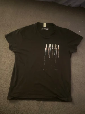 Svart AMIRI t-shirt med tryck - Säljer en svart t-shirt från AMIRI med färgstarkt tryck framtill och baktill där AMIRI-loggan syns med droppande färgeffekt i vitt, blått och orange. T-shirten har korta ärmar och är gjord i mjuk bomull. Perfekt för dig som gillar streetwear och vill sticka ut.